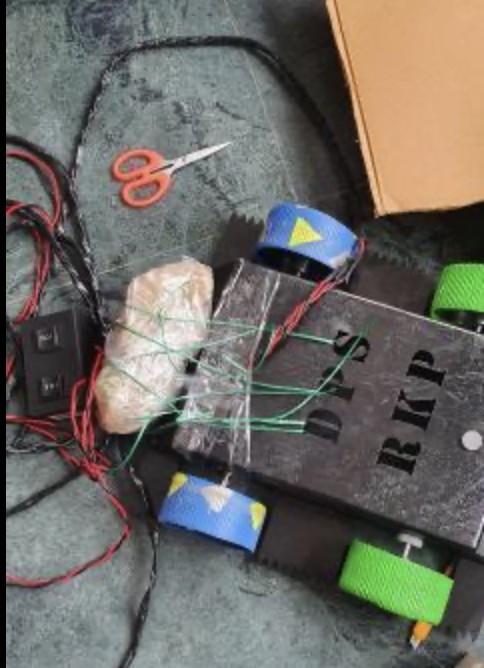 a small black robot with blue and green wheels, a brick strapped to its front with green wire